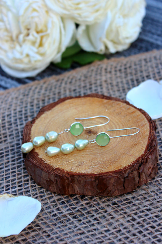 Pair of earrings with green beads and pearls on a wooden slice with white flowers in the background.
