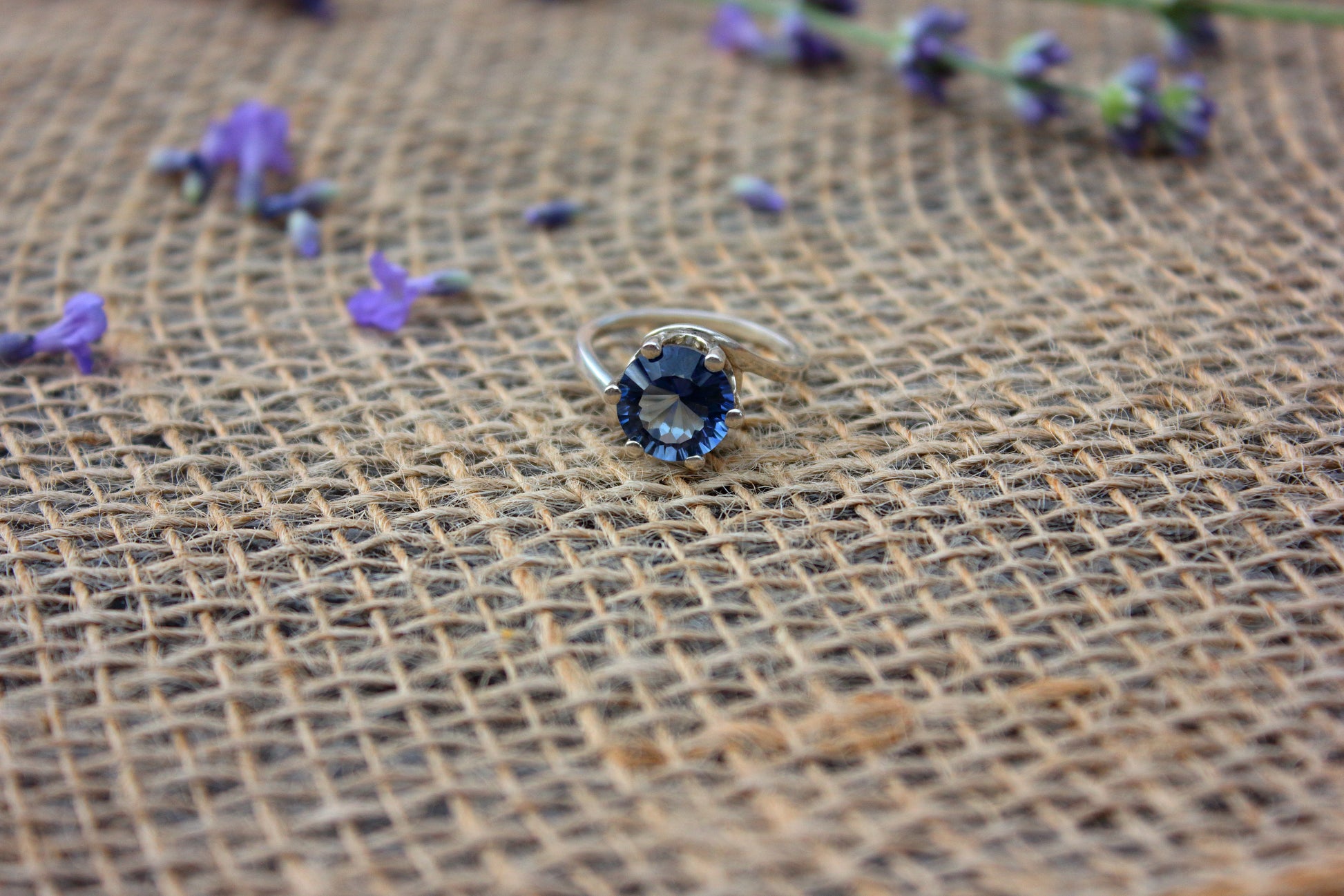 Silver ring with blue gemstone on a textured brown fabric background with scattered purple flowers.