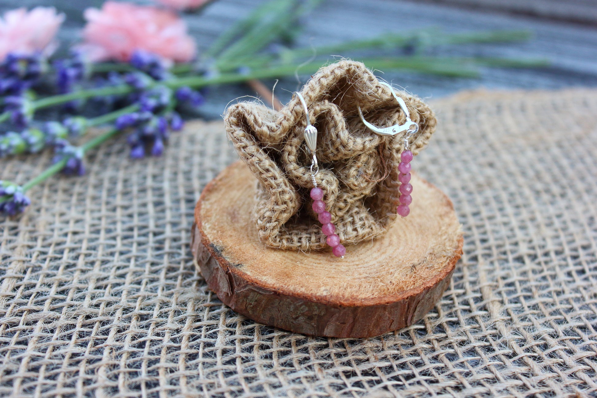 Pair of earrings with pink beads in a decorative woven holder on a wooden surface with flowers in the background.