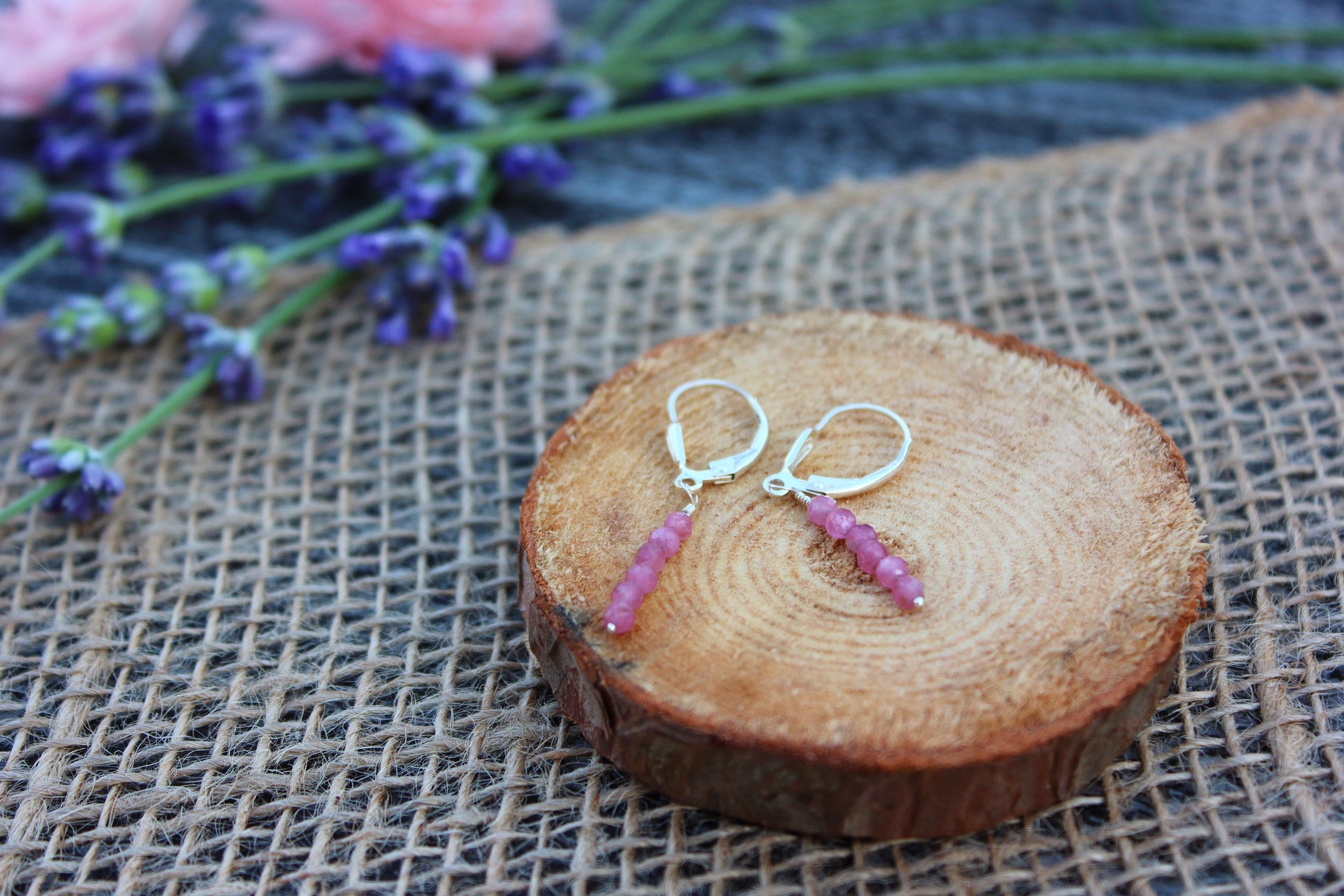 Pair of pink gemstone earrings on a wooden slice with lavender flowers in the background
