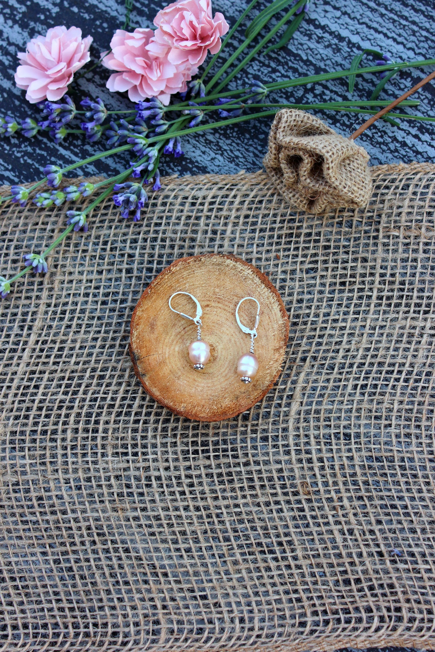 Pair of silver earrings with pink pearls on a wooden coaster, surrounded by pink flowers and lavender.