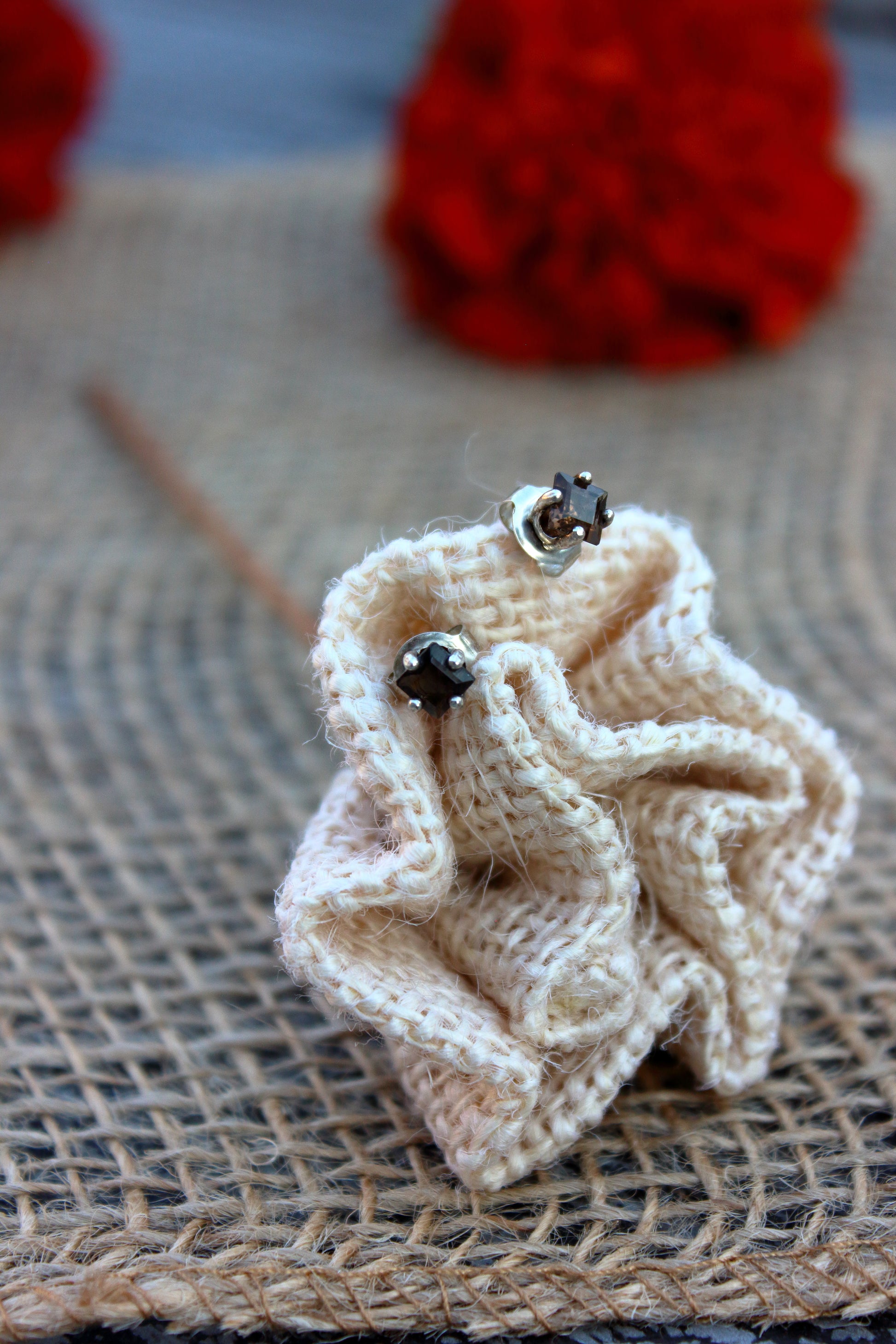 Knitted beige flower and smoky quartz studs on a textured surface with blurred red flowers in the background