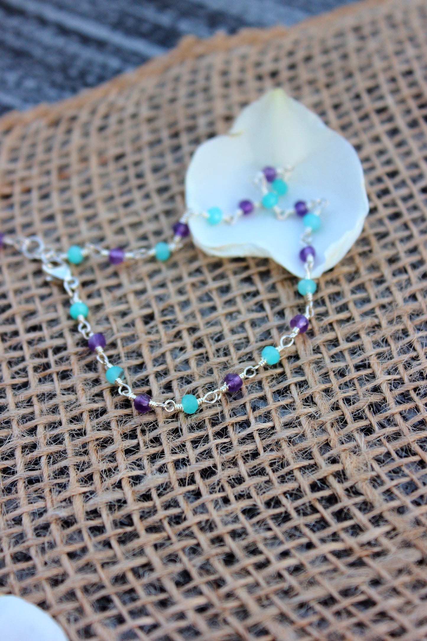 Bracelet with turquoise and purple beads on a white rose petal against a textured brown background