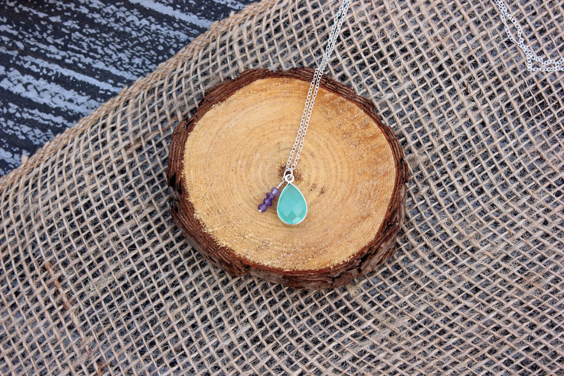Necklace with a teal pendant and purple charm on a wooden stump with burlap background
