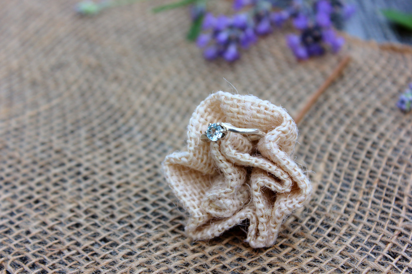 Ring on a knitted beige flower on a textured brown background with blurred purple flowers.