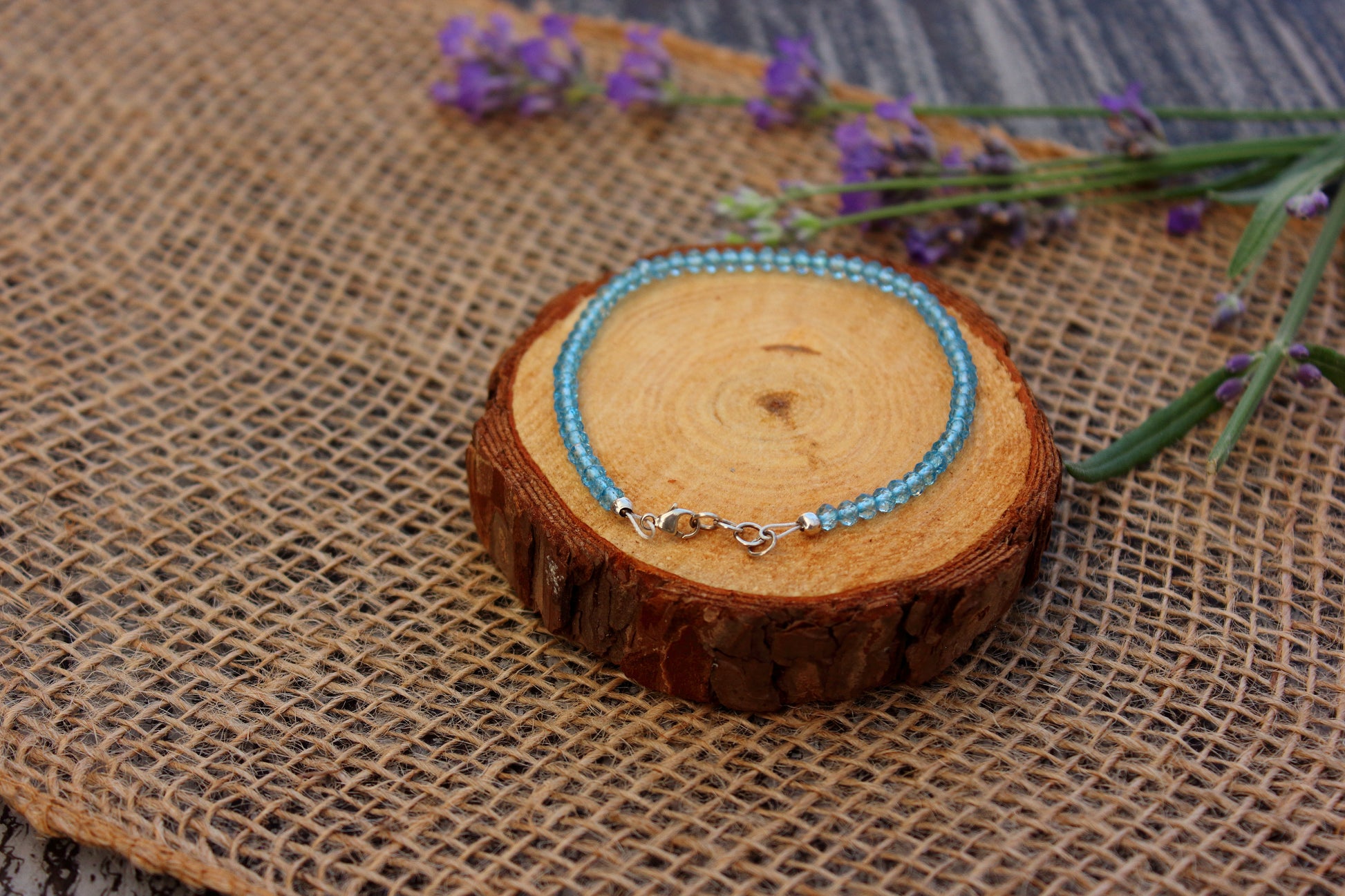 Blue beaded bracelet on a wooden slice with lavender flowers on a textured fabric background
