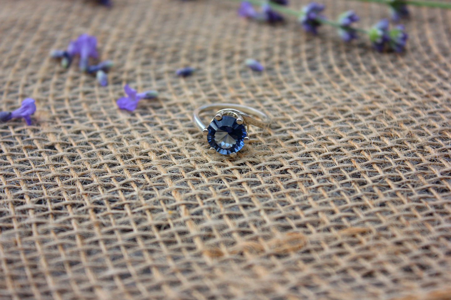 Silver ring with blue gemstone on a textured brown fabric background with scattered purple flowers.