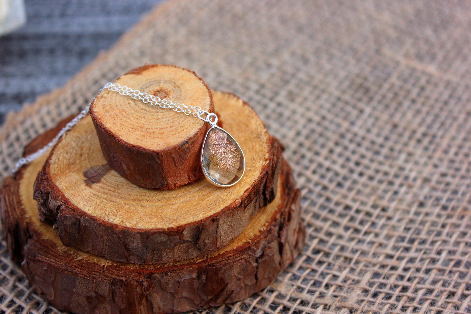 Teardrop-shaped pendant on a chain displayed on wooden slices with a textured fabric background