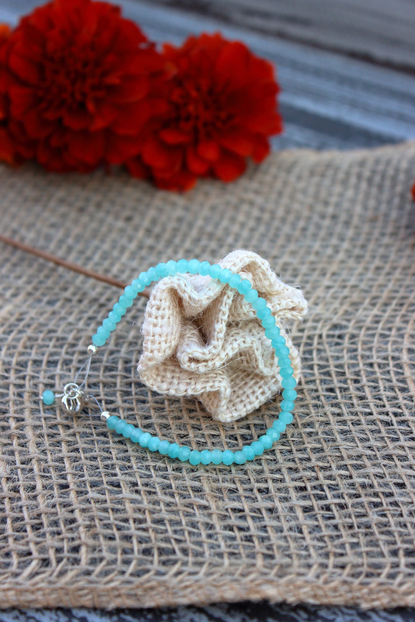 Turquoise beaded bracelet with a beige fabric flower on a textured surface with red flowers in the background.
