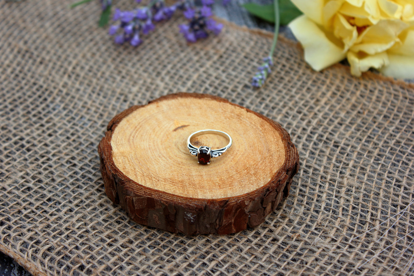Silver ring with a red gemstone on a wooden slice with flowers in the background