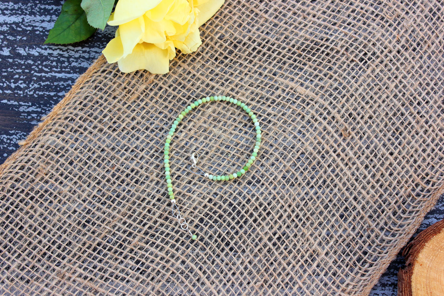 Green beaded bracelet on a textured brown surface with yellow flowers and green leaves.