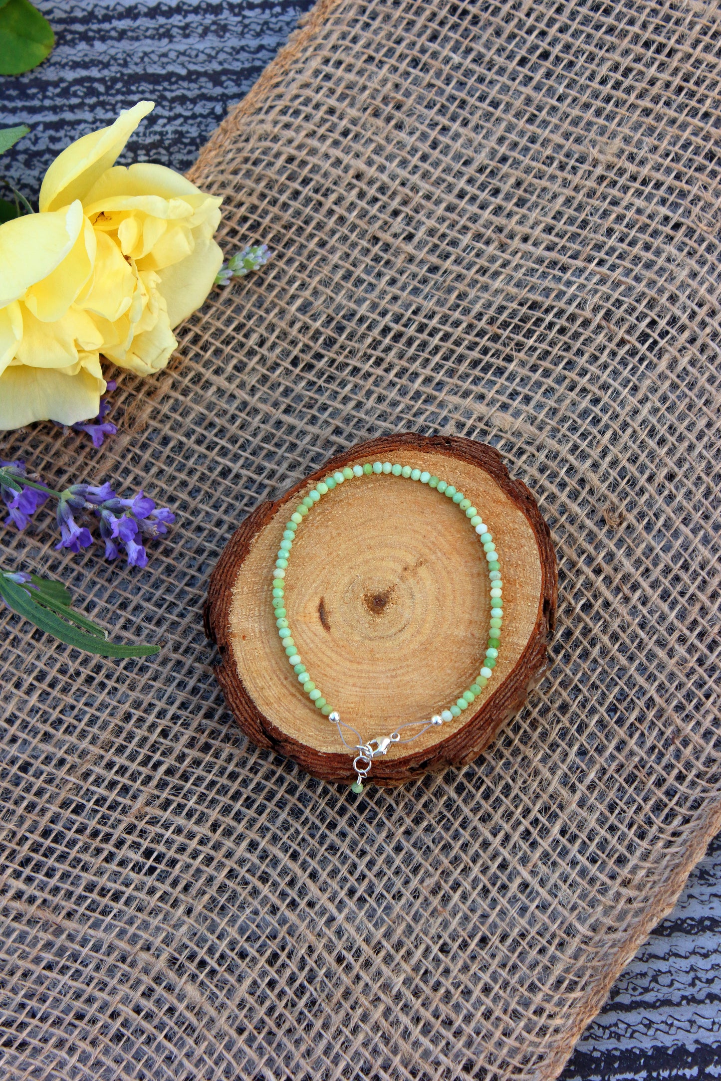 Green beaded bracelet on a wooden slice with yellow flowers and purple lavender on a textured fabric background
