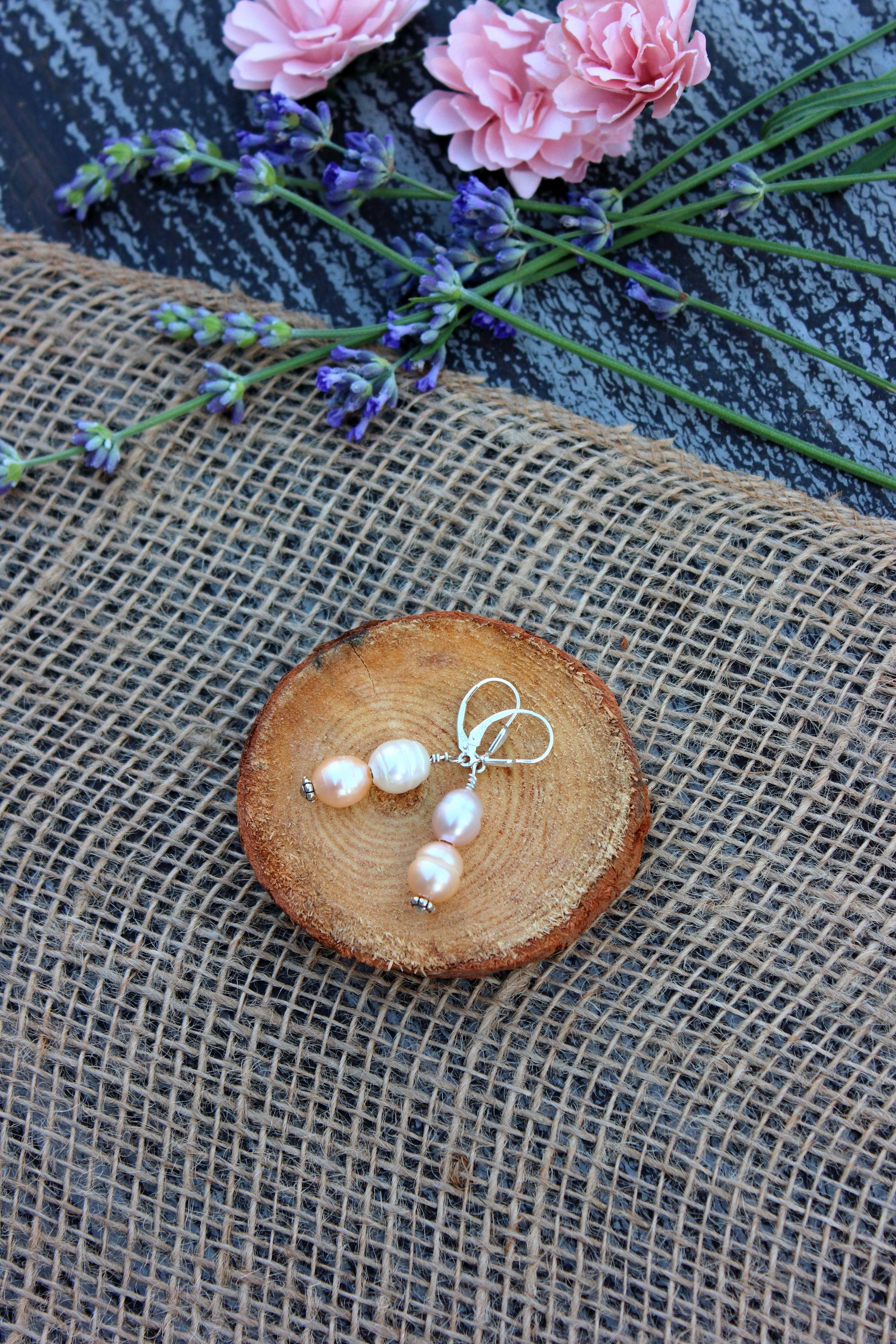 Pearl earrings on a wooden slice with pink flowers and lavender in the background
