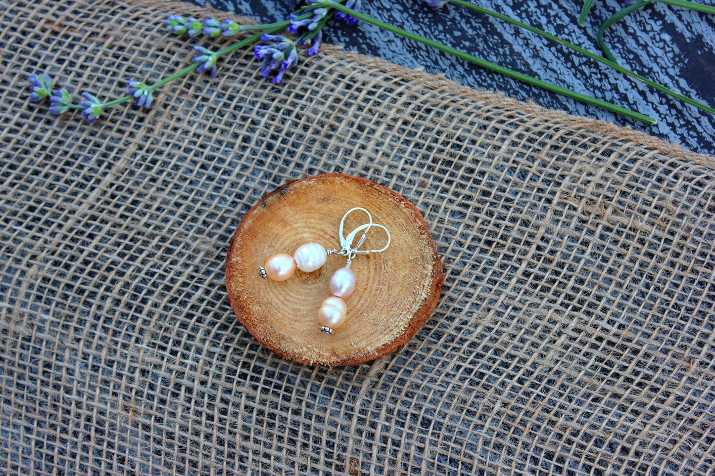 Pearl earrings on a wooden slice with lavender flowers on a textured fabric background