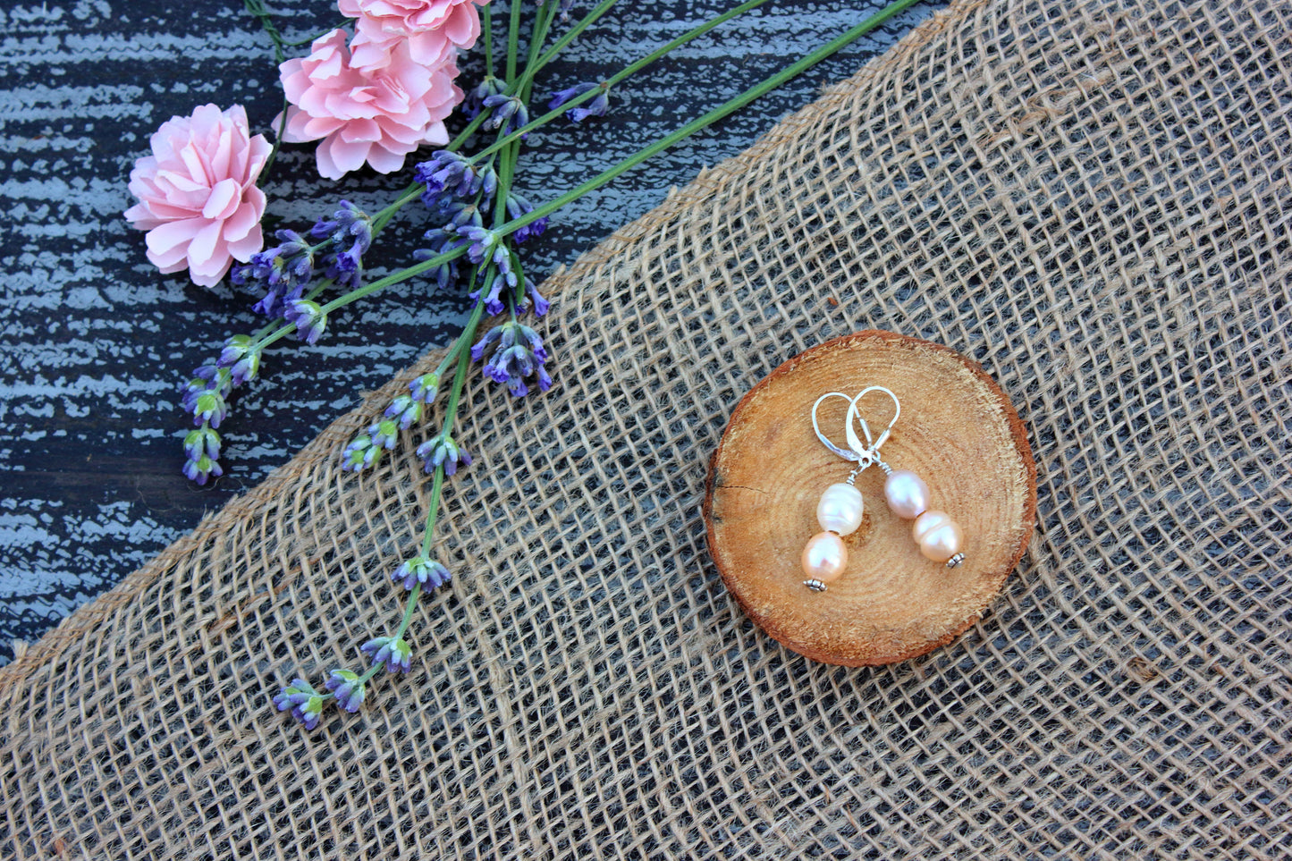 Pearl earrings on a wooden slice with pink flowers and lavender on burlap and denim background