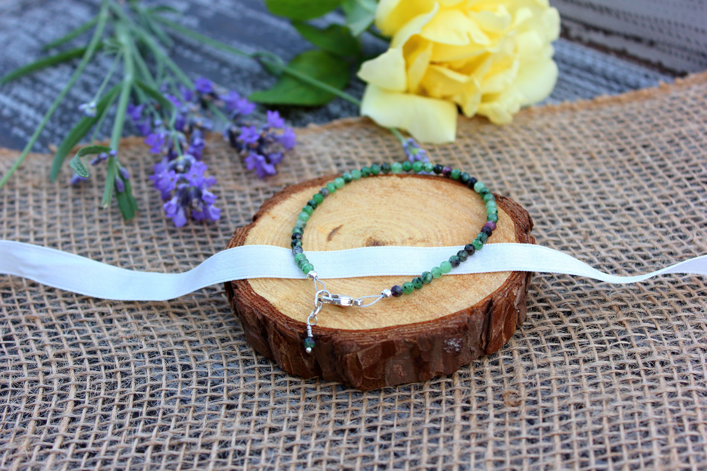 Beaded bracelets on a wooden slice with a white ribbon, surrounded by lavender and a yellow flower on a textured fabric background.