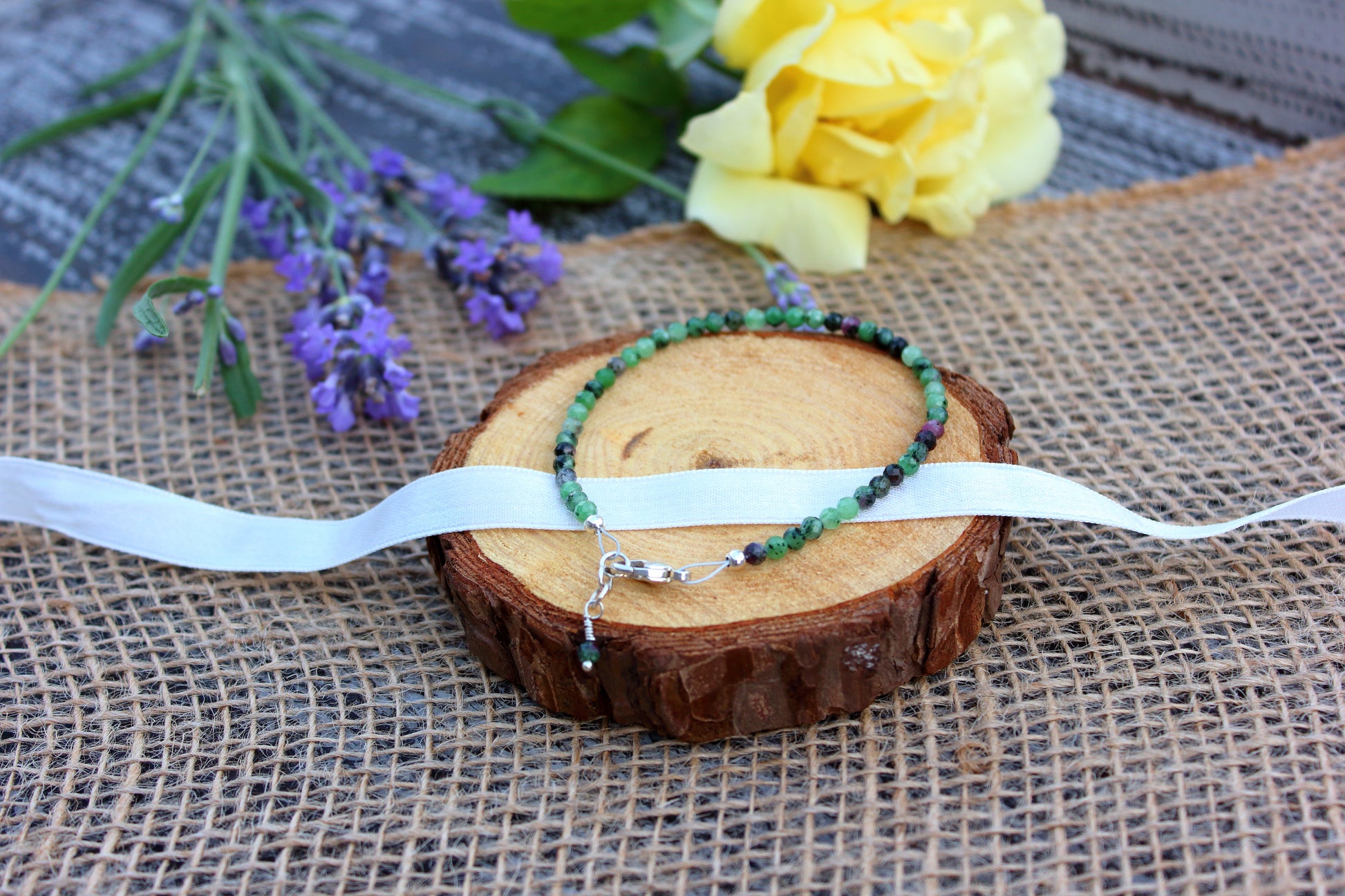 Beaded bracelets on a wooden slice with a white ribbon, surrounded by lavender and a yellow flower on a textured fabric background.
