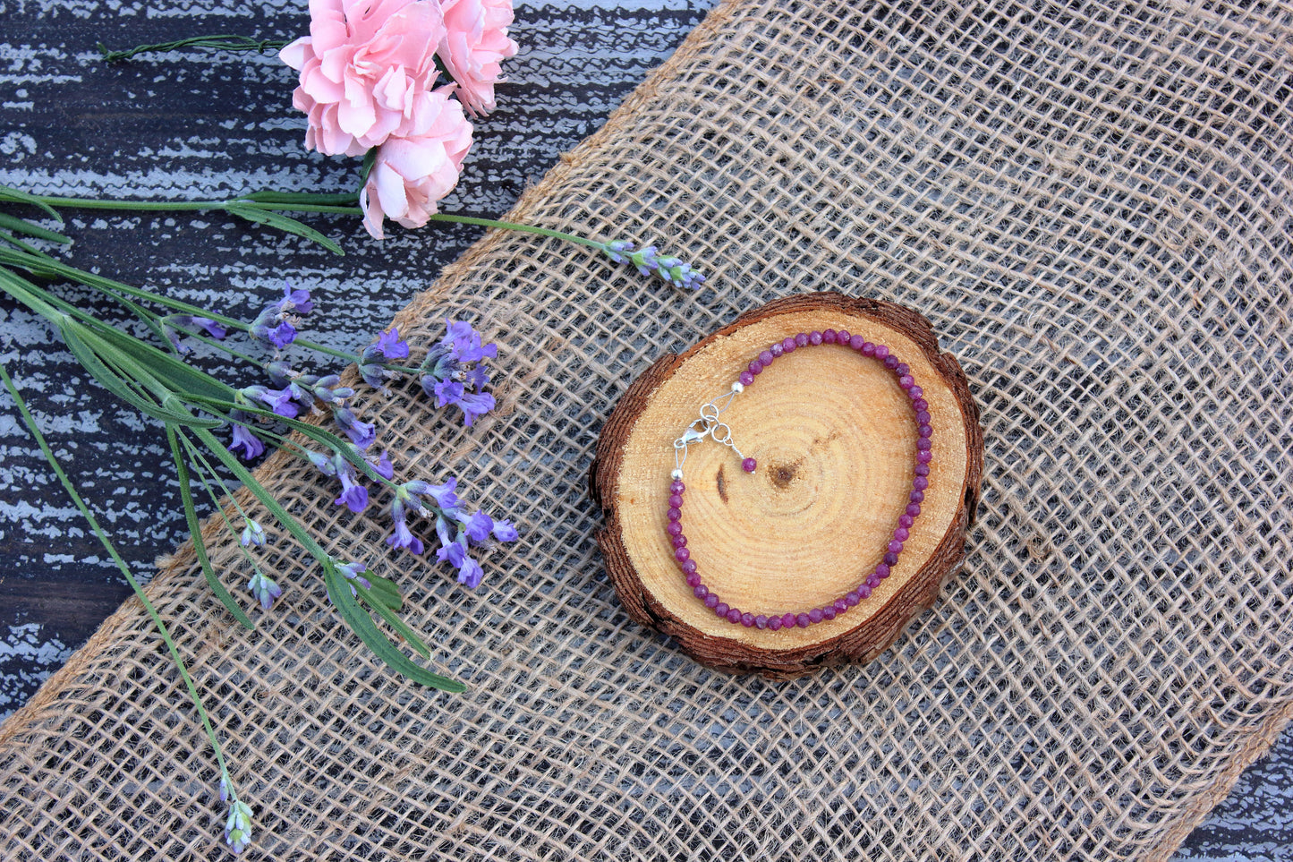 Ruby beaded bracelet on a wooden slice with pink flowers and lavender on a textured surface