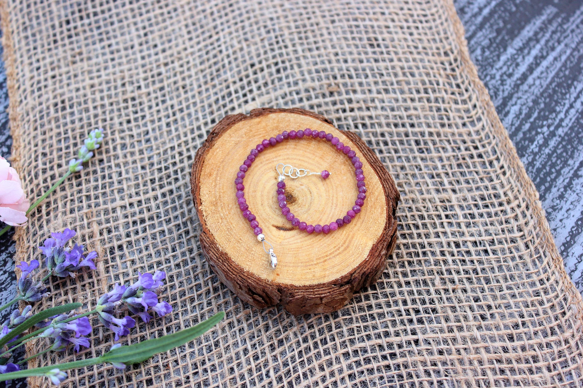 Ruby beaded bracelet on a wooden disc with lavender flowers on a textured fabric background